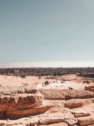 A vast desert landscape with sandy terrain stretching into the distance. In the foreground, large stone blocks hint at ancient ruins. Several camels and people are scattered across the area, with a bustling city skyline visible in the background under a clear blue sky.