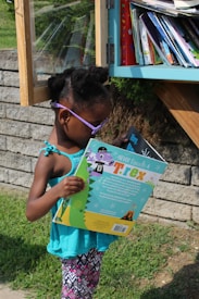 A child wearing purple sunglasses and a turquoise tank top is reading a colorful book titled 'Never Touch a T. rex' near a small outdoor library box. The library is filled with various books and is set against a stone wall in a grassy area.