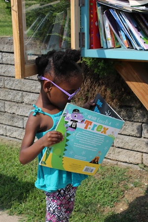 A child wearing purple sunglasses and a turquoise tank top is reading a colorful book titled 'Never Touch a T. rex' near a small outdoor library box. The library is filled with various books and is set against a stone wall in a grassy area.