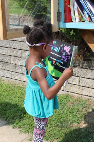 A young child standing outdoors in front of a small library box, intently reading a colorful children's book. She wears a turquoise sleeveless top and patterned leggings with her hair styled in two small puffs. In the background, books are visible in the library box, and the setting is a sunny day with grass and a brick wall nearby.