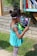 A young child standing outdoors in front of a small library box, intently reading a colorful children's book. She wears a turquoise sleeveless top and patterned leggings with her hair styled in two small puffs. In the background, books are visible in the library box, and the setting is a sunny day with grass and a brick wall nearby.