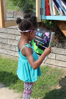 A young child standing outdoors in front of a small library box, intently reading a colorful children's book. She wears a turquoise sleeveless top and patterned leggings with her hair styled in two small puffs. In the background, books are visible in the library box, and the setting is a sunny day with grass and a brick wall nearby.