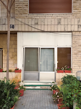 A front entrance with glass sliding doors surrounded by brick walls. The area is decorated with potted plants and flowers on either side of the entrance. An uneven brick path leads up to the door, with foliage creating a welcoming atmosphere. A shuttered window is above the doorway.