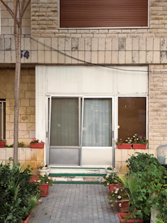 A front entrance with glass sliding doors surrounded by brick walls. The area is decorated with potted plants and flowers on either side of the entrance. An uneven brick path leads up to the door, with foliage creating a welcoming atmosphere. A shuttered window is above the doorway.