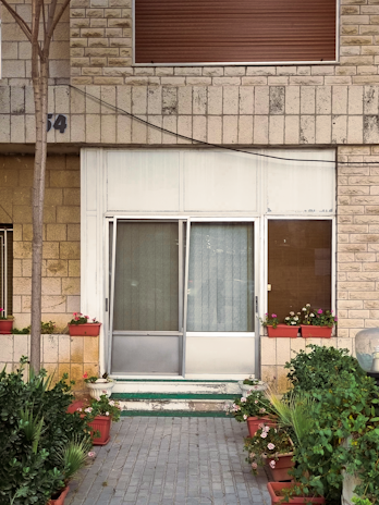 A front entrance with glass sliding doors surrounded by brick walls. The area is decorated with potted plants and flowers on either side of the entrance. An uneven brick path leads up to the door, with foliage creating a welcoming atmosphere. A shuttered window is above the doorway.