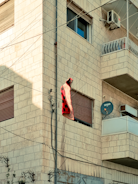 A building made of light-colored bricks featuring multiple windows, an air conditioning unit, and a balcony with a metal railing. Electrical wires run vertically and horizontally across the building. A red and black checkered blanket or cloth is hanging from the wires near one of the windows. There is also a satellite dish attached to the wall below the balcony.