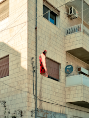 A building made of light-colored bricks featuring multiple windows, an air conditioning unit, and a balcony with a metal railing. Electrical wires run vertically and horizontally across the building. A red and black checkered blanket or cloth is hanging from the wires near one of the windows. There is also a satellite dish attached to the wall below the balcony.