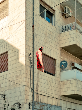A building made of light-colored bricks featuring multiple windows, an air conditioning unit, and a balcony with a metal railing. Electrical wires run vertically and horizontally across the building. A red and black checkered blanket or cloth is hanging from the wires near one of the windows. There is also a satellite dish attached to the wall below the balcony.