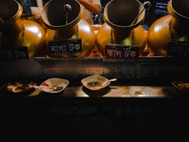 Four large brass containers with labels in a non-English script are arranged side by side, each with a ladle sticking out. In front of them on a counter are three used plates and spoons with remnants of food, possibly indicating a street food or beverage setup.