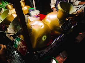 A collection of large, shiny, brass vessels is arranged in a display. Each vessel has a wide, open mouth and is connected to a board with labels in a script that isn't Latin. In the background, a person is partially visible, wearing a light-colored shirt, and appears to be involved in a food or drink preparation process. The setting suggests a street food stall with various utensils and containers.