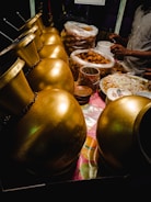 A row of shiny, golden urns is placed alongside a table filled with various food items, including stacks of fried snacks in plastic bags and bowls. A person is partially visible, busy preparing or serving food, with their hands in motion.