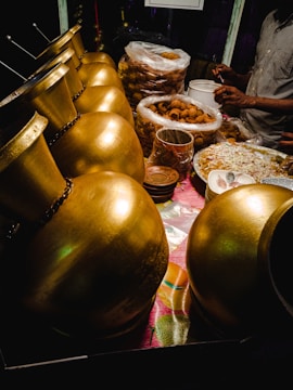 A row of shiny, golden urns is placed alongside a table filled with various food items, including stacks of fried snacks in plastic bags and bowls. A person is partially visible, busy preparing or serving food, with their hands in motion.