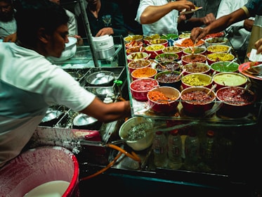A street food vendor is serving a variety of colorful dishes from multiple pots and bowls. The setup is bustling with a mix of sauces, spices, and condiments, each offering vibrant colors like red, green, and yellow. Several people are involved in the preparation and serving of the food.