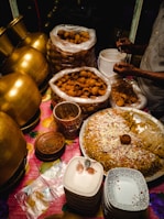 A food stall setup featuring large brass pots, stacks of ceramic plates, and a variety of Indian street food items, including a large tray of seasoned rice garnished with onions and spices. Plastic bags full of crispy snacks are placed nearby, with a person serving them into smaller containers.