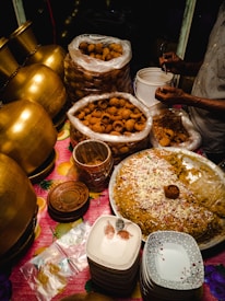 A food stall setup featuring large brass pots, stacks of ceramic plates, and a variety of Indian street food items, including a large tray of seasoned rice garnished with onions and spices. Plastic bags full of crispy snacks are placed nearby, with a person serving them into smaller containers.