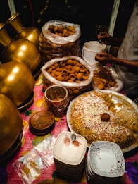 A food stall setup featuring large brass pots, stacks of ceramic plates, and a variety of Indian street food items, including a large tray of seasoned rice garnished with onions and spices. Plastic bags full of crispy snacks are placed nearby, with a person serving them into smaller containers.