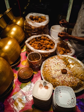 A food stall setup featuring large brass pots, stacks of ceramic plates, and a variety of Indian street food items, including a large tray of seasoned rice garnished with onions and spices. Plastic bags full of crispy snacks are placed nearby, with a person serving them into smaller containers.