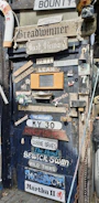 A rustic door is covered with a variety of signs and plaques displaying different names and phrases. Items such as a life ring, nautical decorations, and various vintage memorabilia are attached around the door. A mix of wooden and metal objects, including a chain and lantern, contribute to the eclectic and nautical theme.