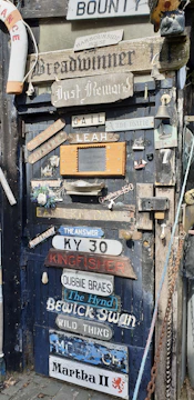 A rustic door is covered with a variety of signs and plaques displaying different names and phrases. Items such as a life ring, nautical decorations, and various vintage memorabilia are attached around the door. A mix of wooden and metal objects, including a chain and lantern, contribute to the eclectic and nautical theme.