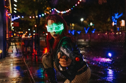 A person with neon green light glasses and red hair crouches on a wet, reflective street at night. They are wearing a black leather jacket and fishnet stockings, pointing a futuristic toy gun towards the camera. Illuminated colorful string lights and blurred city lights create a cyberpunk atmosphere.