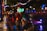 A person with neon green light glasses and red hair crouches on a wet, reflective street at night. They are wearing a black leather jacket and fishnet stockings, pointing a futuristic toy gun towards the camera. Illuminated colorful string lights and blurred city lights create a cyberpunk atmosphere.