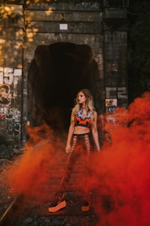 A person stands confidently on a railway track with vibrant red smoke swirling around. The scene is framed by a dark tunnel that's partially covered in graffiti. The outfit includes striking lace-up leggings and platform shoes, adding a bold fashion statement to the mysterious and edgy ambiance.