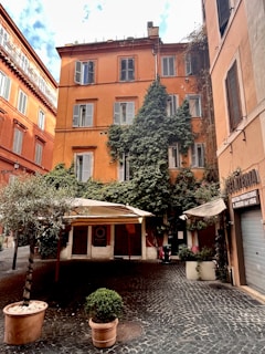 A rustic building with a warm orange facade, adorned with green ivy climbing up one side. It is located in a small courtyard paved with cobblestones, featuring potted plants and an outdoor seating area covered with canvas canopies. The surroundings give a cozy and charming atmosphere.