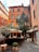 A rustic building with a warm orange facade, adorned with green ivy climbing up one side. It is located in a small courtyard paved with cobblestones, featuring potted plants and an outdoor seating area covered with canvas canopies. The surroundings give a cozy and charming atmosphere.