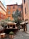 A rustic building with a warm orange facade, adorned with green ivy climbing up one side. It is located in a small courtyard paved with cobblestones, featuring potted plants and an outdoor seating area covered with canvas canopies. The surroundings give a cozy and charming atmosphere.