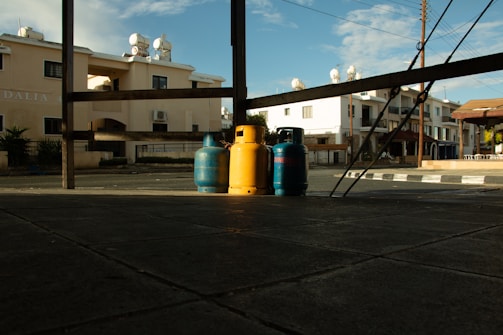 Three gas cylinders, one yellow and two blue, are placed on a tiled pavement under a wooden structure. The background features residential buildings with balconies and satellite dishes, suggesting an urban setting. The sky is partially cloudy, and the lighting suggests late afternoon or early morning.