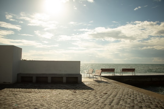 A sunlit seascape with two empty wooden benches facing the ocean. The scene includes a cobblestone pathway and a flat white wall. The sky is partly cloudy, creating soft shadows and a tranquil atmosphere.