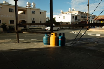 Three gas cylinders, two blue and one yellow, are positioned on a paved street corner under a wooden structure. Residential buildings with solar water heaters on the roofline are visible in the background. A white car is parked across the street, and the scene is partially shaded, indicating a time close to sunset or sunrise.