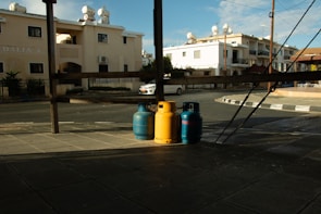Three gas cylinders, two blue and one yellow, are positioned on a paved street corner under a wooden structure. Residential buildings with solar water heaters on the roofline are visible in the background. A white car is parked across the street, and the scene is partially shaded, indicating a time close to sunset or sunrise.