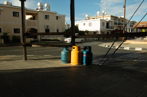 Three gas cylinders, two blue and one yellow, are positioned on a paved street corner under a wooden structure. Residential buildings with solar water heaters on the roofline are visible in the background. A white car is parked across the street, and the scene is partially shaded, indicating a time close to sunset or sunrise.