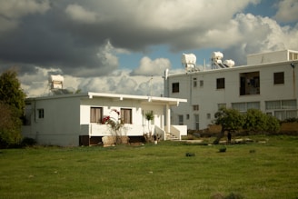 A small white house is flanked by a larger building on the right, both with solar heaters on their roofs. The front of the house features flowering plants, while the lawn in the foreground is vibrant green. The sky is gray and overcast with thick clouds.