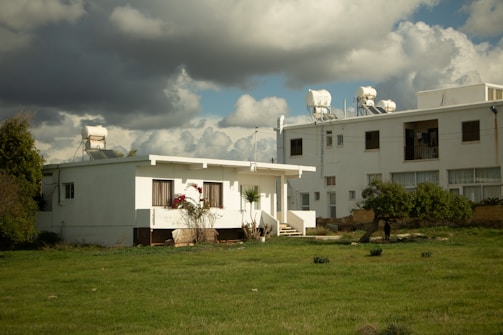 A small white house is flanked by a larger building on the right, both with solar heaters on their roofs. The front of the house features flowering plants, while the lawn in the foreground is vibrant green. The sky is gray and overcast with thick clouds.