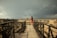 A woman wearing a red dress and blue backpack walks alone along a wooden walkway surrounded by ancient ruins. The sky is overcast with dark storm clouds looming in the distance, creating a dramatic contrast against the earthy tones of the ruins.