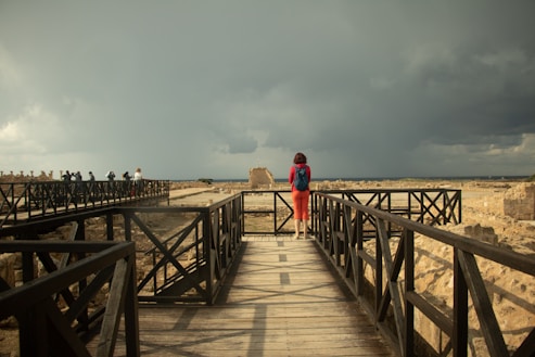 A woman wearing a red dress and blue backpack walks alone along a wooden walkway surrounded by ancient ruins. The sky is overcast with dark storm clouds looming in the distance, creating a dramatic contrast against the earthy tones of the ruins.