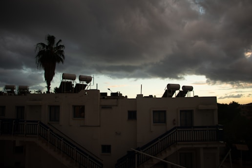 The image captures a white building with external staircases, having solar water heaters mounted on the rooftop. A tall palm tree stands next to the building, silhouetted against a dramatic, cloudy sky. The clouds are dark and heavy, suggesting an impending storm, while a strip of light on the horizon hints at a setting or rising sun.