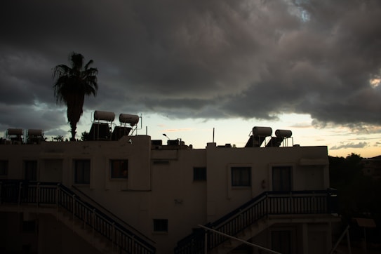 The image captures a white building with external staircases, having solar water heaters mounted on the rooftop. A tall palm tree stands next to the building, silhouetted against a dramatic, cloudy sky. The clouds are dark and heavy, suggesting an impending storm, while a strip of light on the horizon hints at a setting or rising sun.
