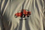 A T-shirt featuring a graphic of a red car with the text 'VW Golf GTI Mk1' printed below the image. The car detailing is bold, with prominent black outlines contributing to a retro aesthetic. The shirt's color is a light, neutral shade.