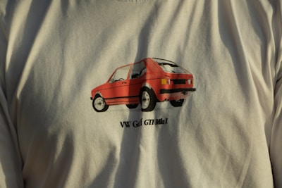 A T-shirt featuring a graphic of a red car with the text 'VW Golf GTI Mk1' printed below the image. The car detailing is bold, with prominent black outlines contributing to a retro aesthetic. The shirt's color is a light, neutral shade.