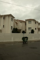 A residential street features creamy white buildings with Mediterranean style architecture, characterized by small balconies and terracotta tiled roofs. A white fence borders the buildings, and a green waste dumpster is placed on the street. The sky appears overcast, casting a subdued light.