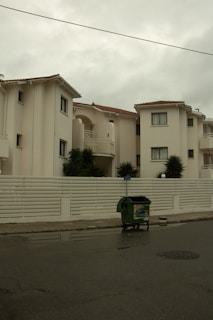 A residential street features creamy white buildings with Mediterranean style architecture, characterized by small balconies and terracotta tiled roofs. A white fence borders the buildings, and a green waste dumpster is placed on the street. The sky appears overcast, casting a subdued light.