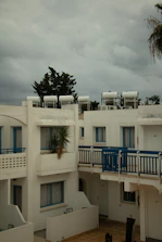 A residential complex with white buildings featuring blue accents, including window frames and railings. Solar water heaters are visible on the rooftops. The sky is overcast with grey clouds, and there is a palm tree partially visible on the right.