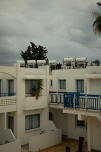 A residential complex with white buildings featuring blue accents, including window frames and railings. Solar water heaters are visible on the rooftops. The sky is overcast with grey clouds, and there is a palm tree partially visible on the right.