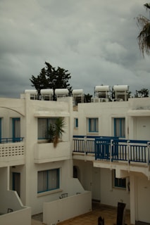 A residential complex with white buildings featuring blue accents, including window frames and railings. Solar water heaters are visible on the rooftops. The sky is overcast with grey clouds, and there is a palm tree partially visible on the right.