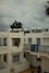 A residential complex with white buildings featuring blue accents, including window frames and railings. Solar water heaters are visible on the rooftops. The sky is overcast with grey clouds, and there is a palm tree partially visible on the right.