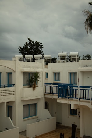 A residential complex with white buildings featuring blue accents, including window frames and railings. Solar water heaters are visible on the rooftops. The sky is overcast with grey clouds, and there is a palm tree partially visible on the right.