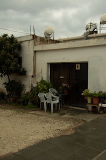 A simple patio area with two white plastic chairs and a small table, surrounded by various potted plants. The area is adjacent to a building with an old and slightly weathered exterior. The rooftop has water tanks and satellite dishes. The ground is a mix of stone tiles and gravel.